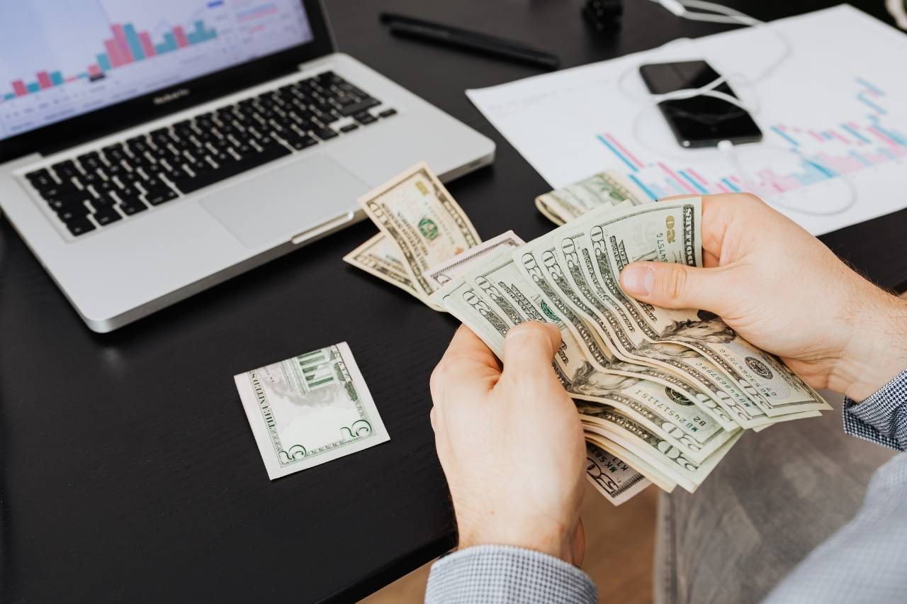 Person counting cash at a desk with a laptop and financial charts in the background.