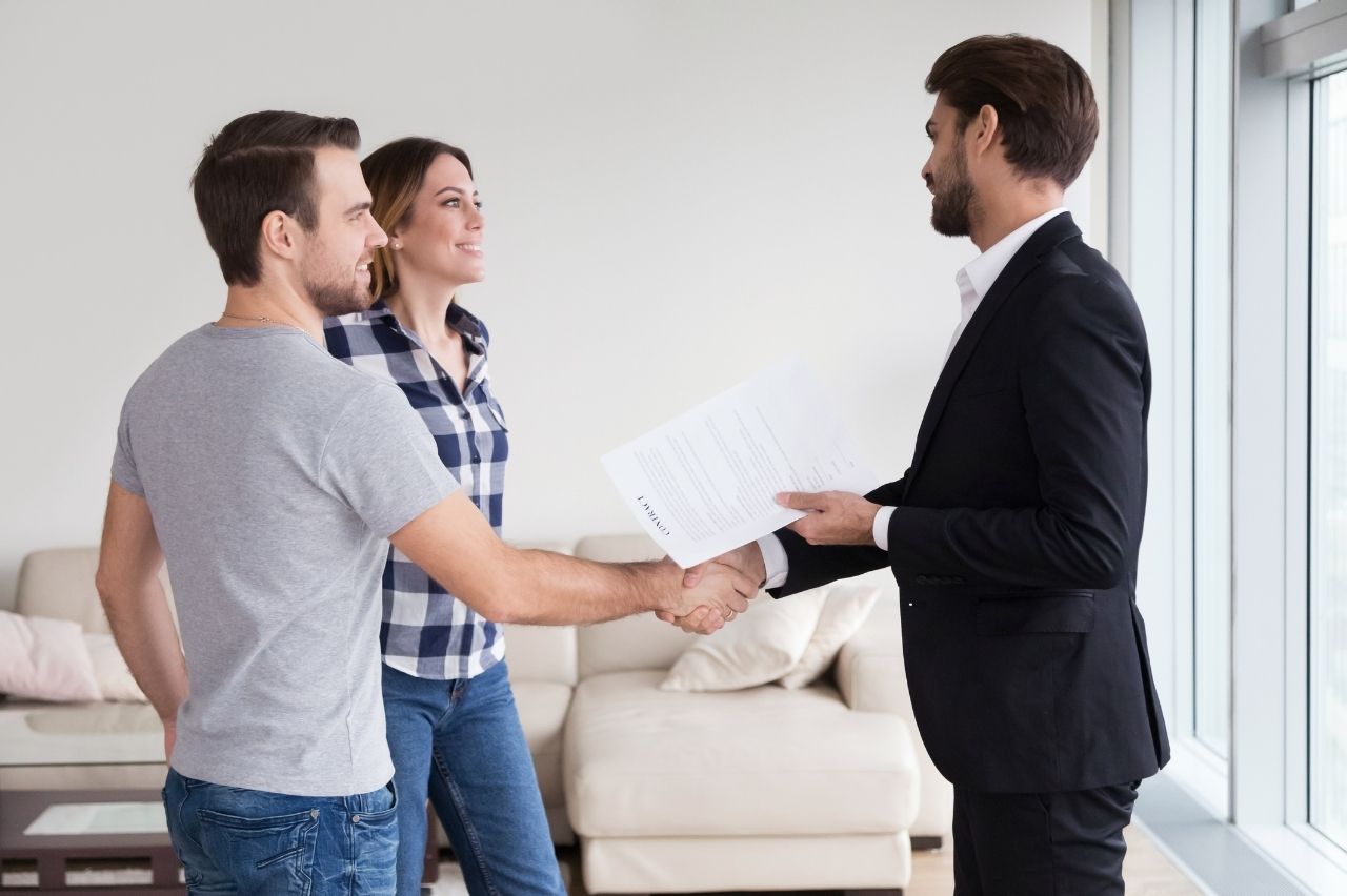 A couple shakes hands with a professional holding a contract inside a bright, modern apartment.